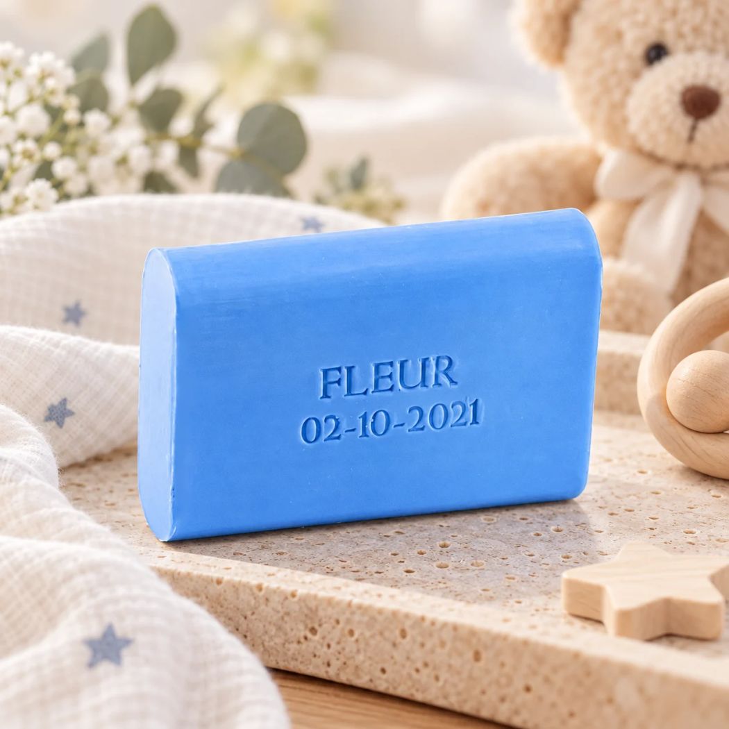 Blauw afgerond zeepje met de naam Fleur en datum 02-10-2021, sfeervol gepresenteerd met zachte stoffen, houten babyaccessoires en een teddybeer op de achtergrond.