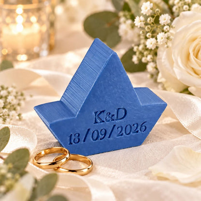 Gepersonaliseerd zeepje in de vorm van een bootje met initialen K&D en trouwdatum 18/09/2026 in de kleur Mure, sfeervol gepresenteerd met witte bloemen en twee gouden trouwringen.