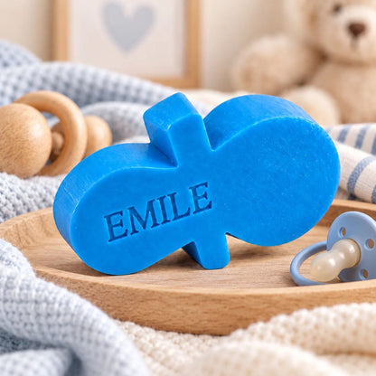 Gepersonaliseerd zeepje in de vorm van een fopspeen met de naam Emile, in de kleur azuur, sfeervol gepresenteerd met blauwe babyaccessoires, houten speelgoed en een blauwe fopspeen.