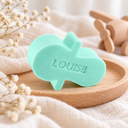 Gepersonaliseerd zeepje in de vorm van een fopspeen met de naam Louise, in de kleur muguet, sfeervol gepresenteerd met zachte babytextielen en houten speelgoed.