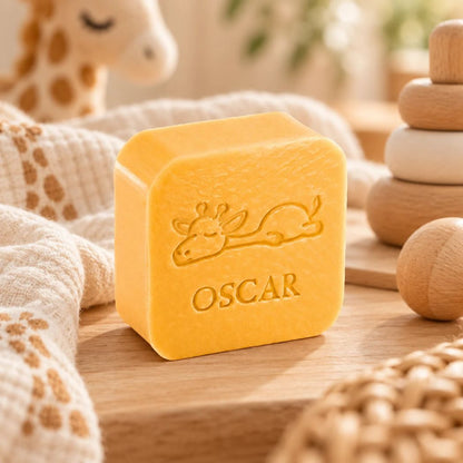 Gepersonaliseerd vierkant zeepje met giraf en de naam Oscar in kleur Kamperfoelie, sfeervol gepresenteerd op een houten ondergrond met zachte stoffen en houten babyaccessoires.