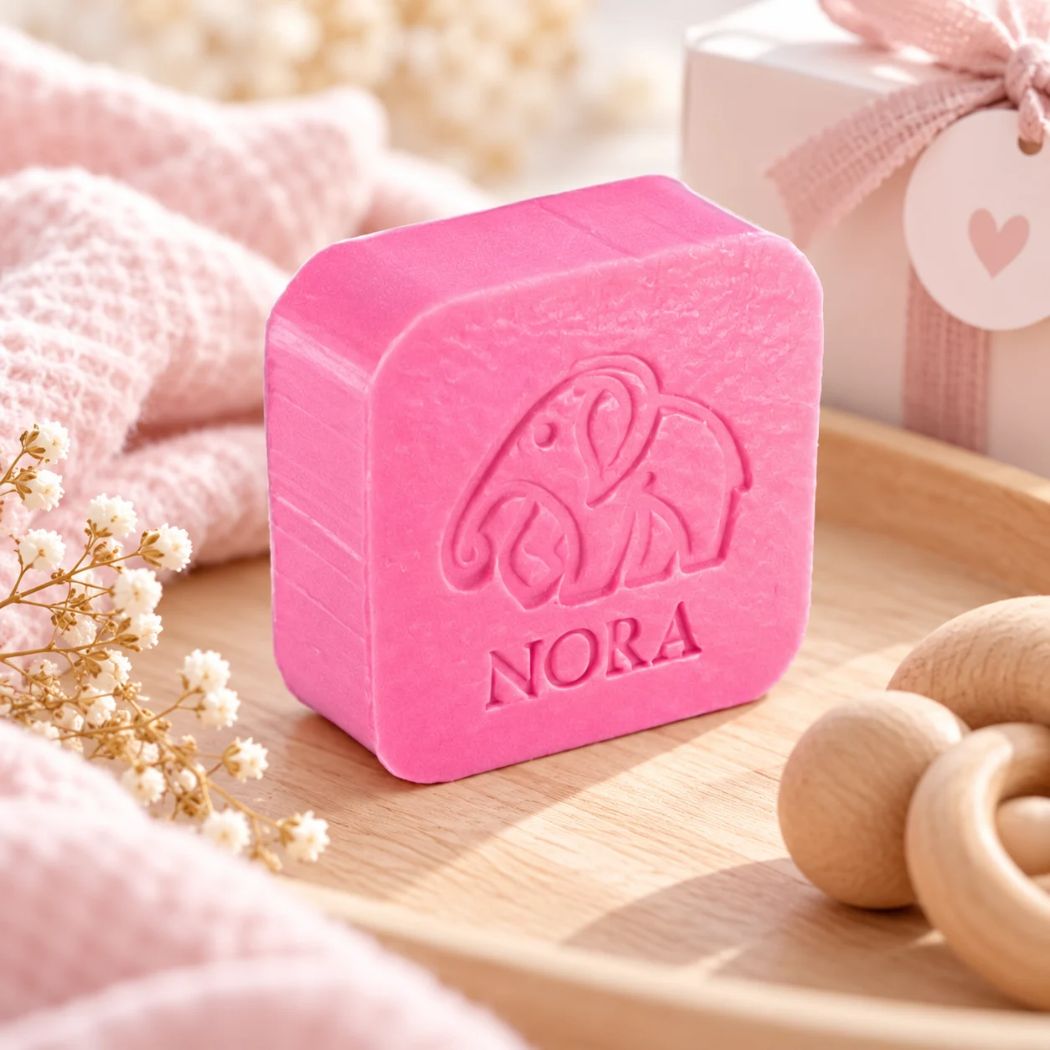 Gepersonaliseerd zeepje met olifantmotief en de naam Nora, in de kleur passion, sfeervol gepresenteerd met roze babytextielen en houten speelgoed.