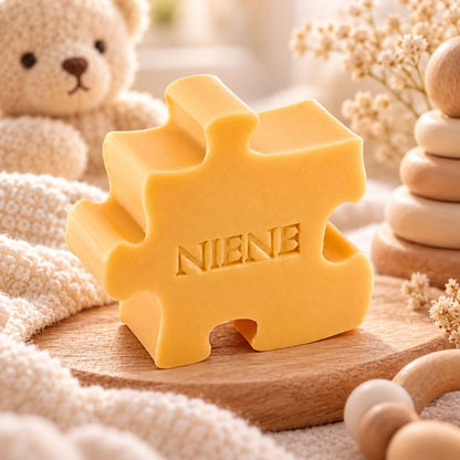 Gepersonaliseerd puzzelzeepje met de naam Niene in kleur Kamperfoelie. Sfeervol gepresenteerd op een houten plankje met zachte stoffen, houten babyaccessoires en een teddybeer op de achtergrond.