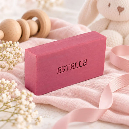 Gepersonaliseerd rechthoekig zeepje met de naam Estelle in de kleur bordo, sfeervol gepresenteerd met roze babytextiel, houten speelgoed en zachte bloemen.
