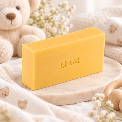 Gepersonaliseerd rechthoekig zeepje met de naam Liam in de kleur kamperfoelie, sfeervol gepresenteerd met zachte babytextielen, teddybeer en houten accessoires.
