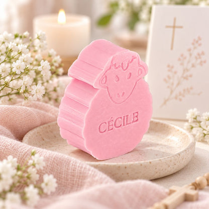 Gepersonaliseerd zeepje in de vorm van een schaap met de naam Cécile, in de kleur rozen, sfeervol gepresenteerd in communiethema met kaars, kruis en zachte witte bloemen.