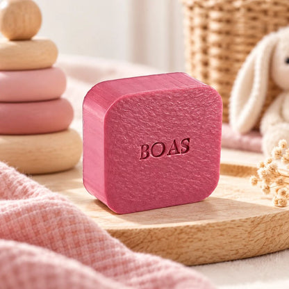 Gepersonaliseerd vierkant zeepje met de naam Boas in kleur Bordo, sfeervol gepresenteerd op een houten schaaltje met zachte roze stoffen en babyaccessoires.