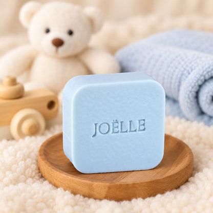 Gepersonaliseerd vierkant zeepje met de naam Joëlle, sfeervol gepresenteerd op een houten schaaltje met zachte babyaccessoires op de achtergrond.
