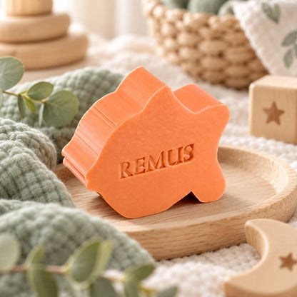 Oranjekleurig zeepje in de vorm van een vis met de naam Remus, sfeervol gepresenteerd op een houten schaaltje met zachtgroene stoffen en houten babyaccessoires.
