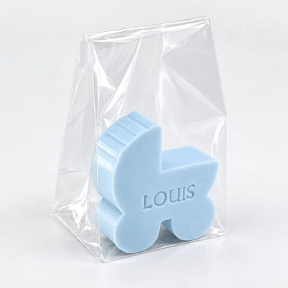 Gepersonaliseerd zeepje in de vorm van een wiegje met de naam Louis, in de kleur pari, verpakt in een transparant zakje op een lichte achtergrond.