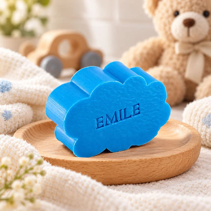 Gepersonaliseerd zeepje in de vorm van een wolk met de naam Emile, in de kleur azuur, sfeervol gepresenteerd met zachte babytextielen, houten speelgoed en een teddybeer.