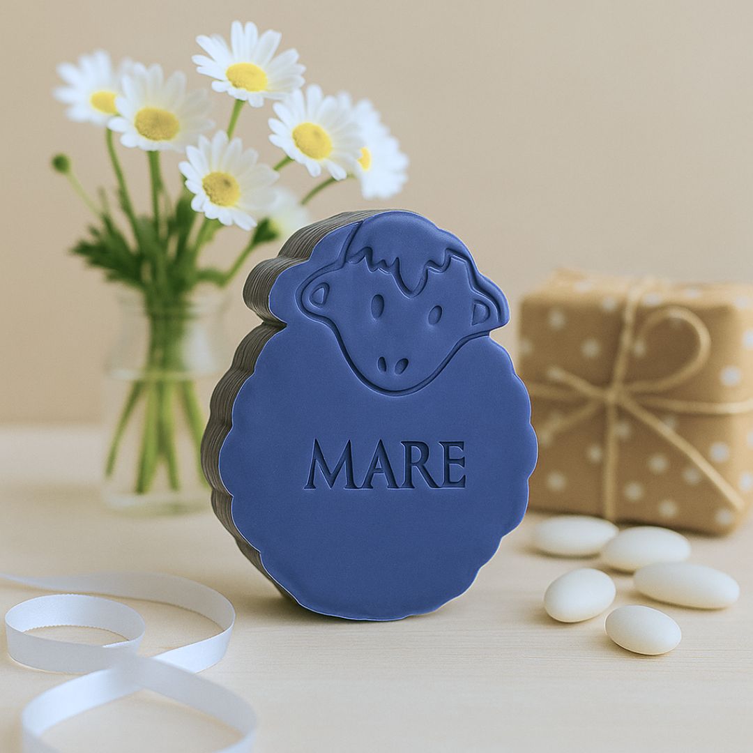 een donkerblauw zeepje in de vorm van een schaap. Op de voorgrond wit lint en suikerbonen. Op de achtergrond een vaas met bloemen en een cadeautje.