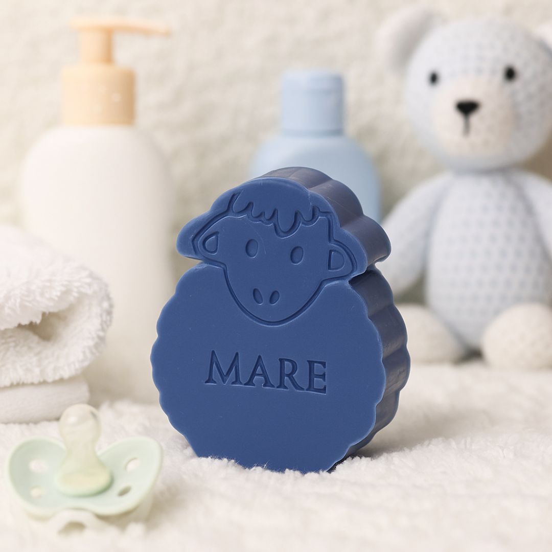 een donkerblauw zeepje in de vorm van een schaap op een schaapsvel. Op de achtergrond een  lichtblauwe teddybeer en op de voorgrond een tutje. 