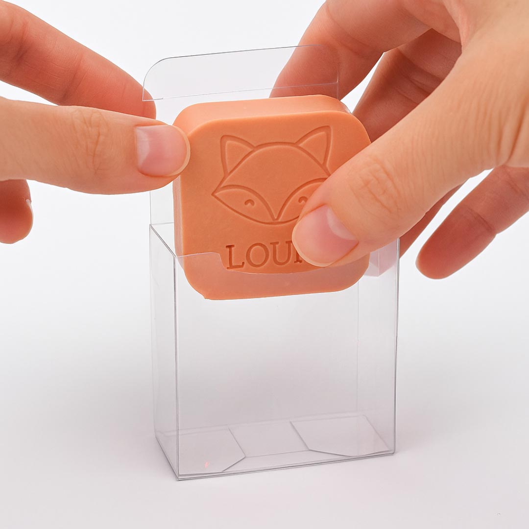 Een hand plaatst een oranje zeepje met vos en naam "LOUIS" in een transparant doosje