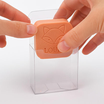 Een hand plaatst een oranje zeepje met vos en naam "LOUIS" in een transparant doosje
